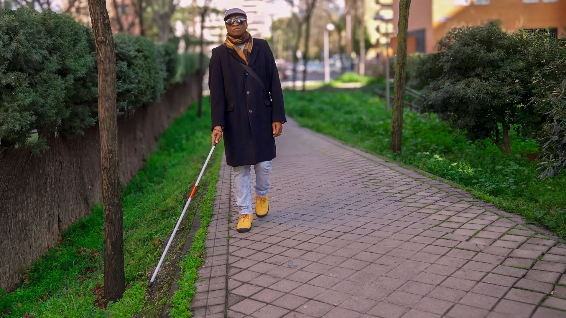 A man in a dark coat and flat cap walks alone along a paved path between hedges, sweeping a long white cane ahead of him.