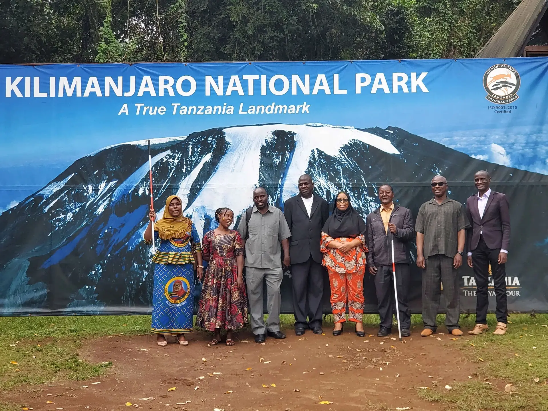 Eight TLB climbers and guides stand in front of the Kilimanjaro National Park banner. Two hold white canes, one raised above her head.