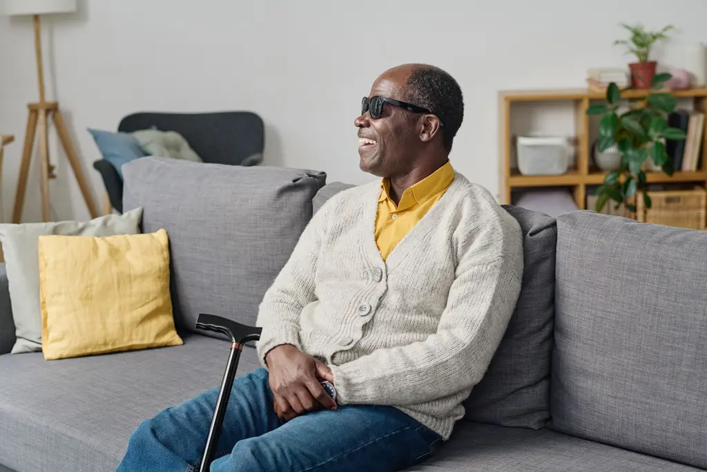 An older man in dark glasses sits back on a sofa at home, smiling, a walking cane resting against his knee.